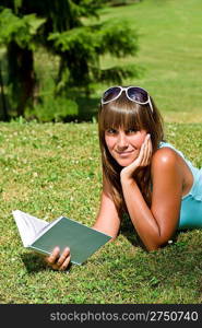 Smiling young woman lying down on grass with book in summer park