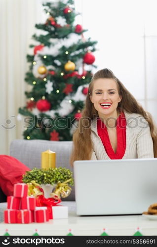 Smiling young woman near Christmas tree sending greeting emails