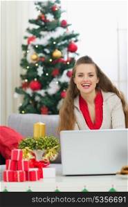 Smiling young woman near Christmas tree sending greeting emails