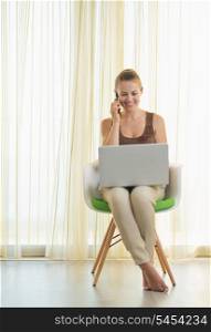 Smiling young woman on modern chair with laptop talking mobile phone