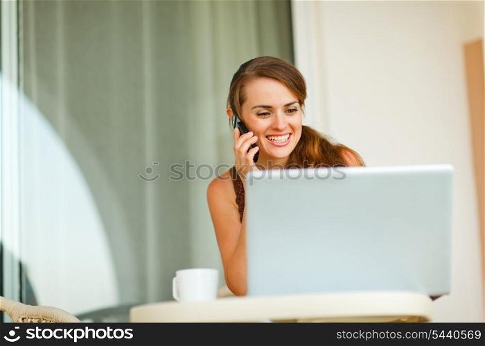 Smiling young woman on terrace speaking cell phone and looking on laptop &#xA;