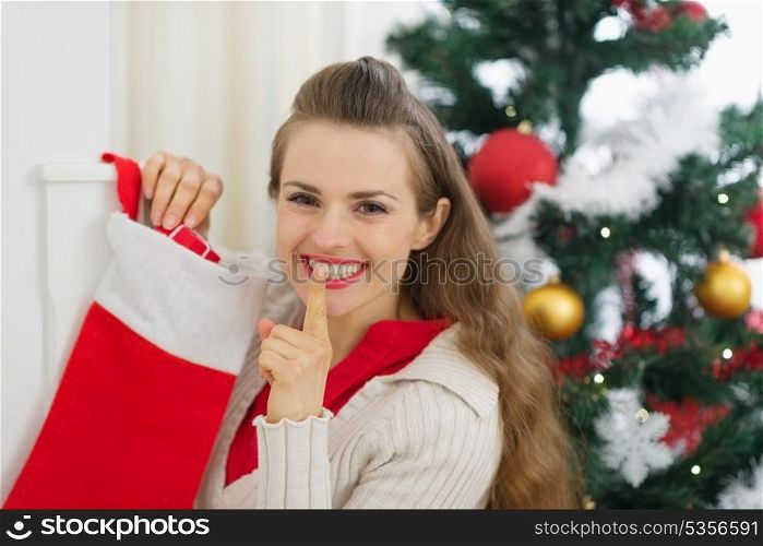 Smiling young woman put gift in Christmas socks and showing shh gesture