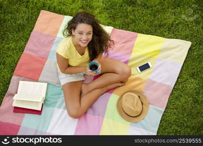 Smiling young woman sitting on a blanket and drinking coffee