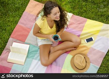 Smiling young woman sitting on a blanket and drinking coffee