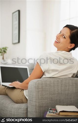 Smiling young woman sitting on couch and working on laptop