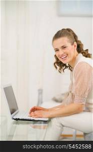 Smiling young woman sitting on couch in living room and using laptop