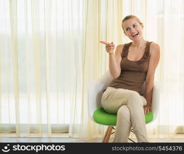 Smiling young woman sitting on modern chair and pointing on copy space