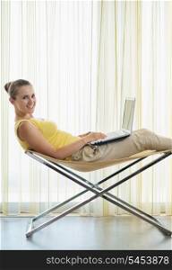 Smiling young woman sitting on modern chair and working on laptop