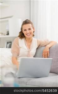 Smiling young woman sitting on sofa in living room with laptop