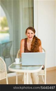 Smiling young woman sitting on terrace with cup of coffee and laptop&#xA;