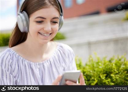 Smiling Young Woman Student On College Campus Streams Music From Mobile Phone To Wireless Headphones