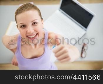 Smiling young woman studying in kitchen and showing thumbs up