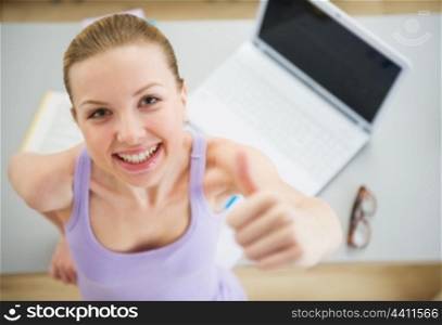 Smiling young woman studying in kitchen and showing thumbs up