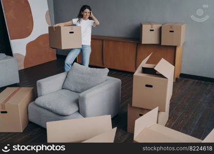 Smiling young woman tenant surrounded by cardboard boxes standing in room moving to new house, happy spanish girl relocates to first own home. Mortgage advertising, relocation concept.. Smiling woman holding cardboard box moving to new home. Mortgage advertising, relocation concept