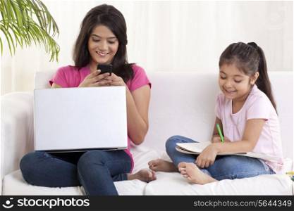 Smiling young woman texting with daughter sitting besides her and drawing