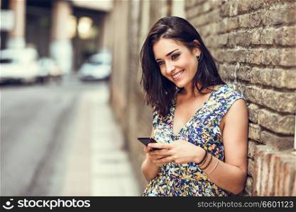 Smiling young woman using her smart phone outdoors. Girl wearing flower dress in urban background. Technology concept.
