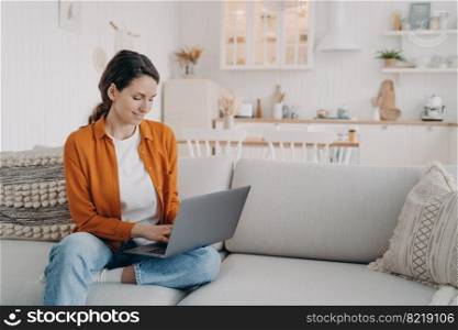 Smiling young woman using laptop, typing email, sitting on comfortable couch, pretty modern girl shopping online, chatting on social networks. Female freelancer working on computer project at home.. Modern girl working at laptop, shopping online, chatting on social networks, sitting on sofa at home