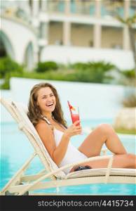 Smiling young woman with cocktail laying on chaise-longue at poolside