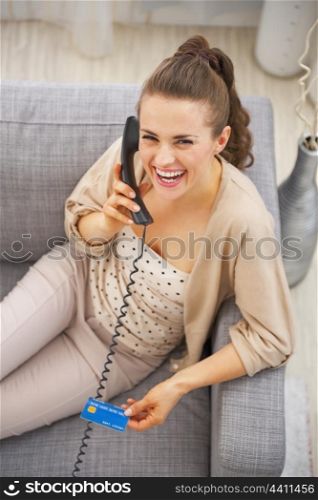 Smiling young woman with credit card sitting on sofa and talking phone