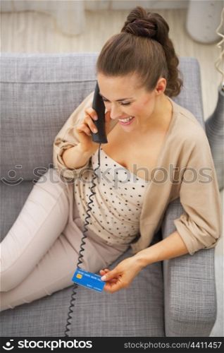 Smiling young woman with credit card sitting on sofa and talking phone