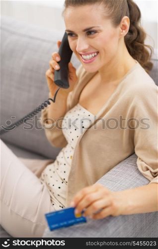 Smiling young woman with credit card talking phone while sitting on sofa