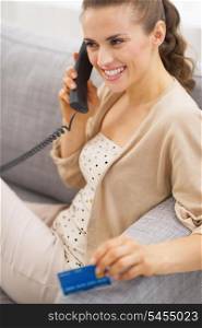Smiling young woman with credit card talking phone while sitting on sofa