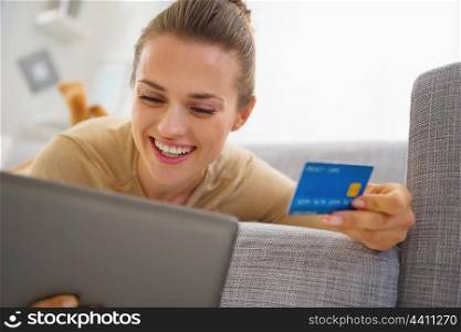 Smiling young woman with credit card using tablet pc while laying on sofa