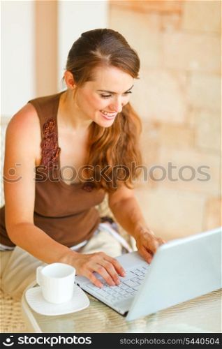 Smiling young woman with cup of coffee working on laptop on terrace &#xA;