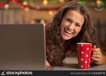 Smiling young woman with cup of hot chocolate looking out from laptop