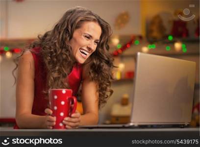 Smiling young woman with cup of hot chocolate usign laptop in christmas decorated kitchen