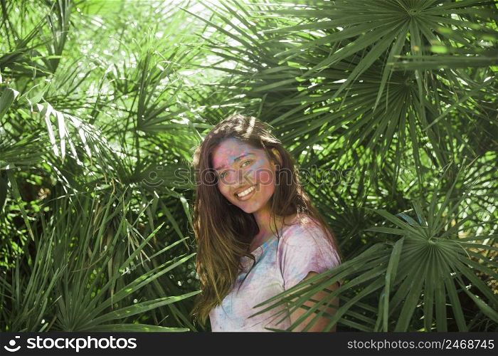 smiling young woman with holi color her body standing among green palm leaves