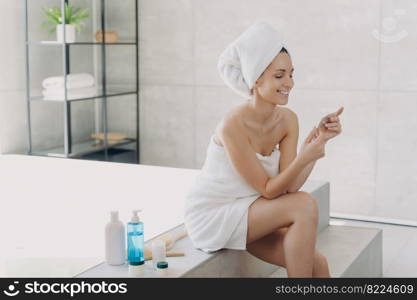 Smiling young woman wrapped in towel reading ingredients list on cosmetics s&le sitting on bathtub in bathroom. Skincare treatment, spa procedure, natural cosmetic products advertising.. Smiling woman wrapped in towel holds cosmetics s&le sitting in bathroom. Skincare treatment, spa