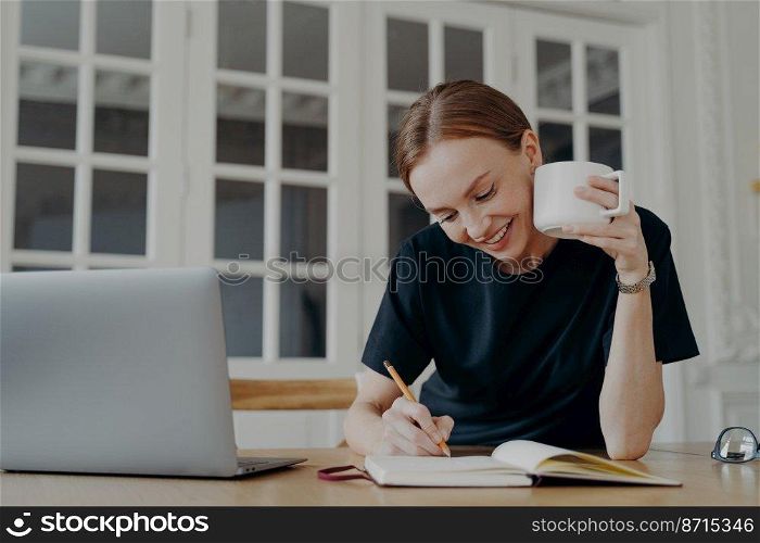 Smiling young woman writing ideas, goals, taking notes, sitting at desk with laptop. Happy freelance female writes in notebook, planning day schedule in the workplace, drinking coffee at home.. Smiling woman writing, taking notes in notebook, planning day schedule, sitting at desk with laptop