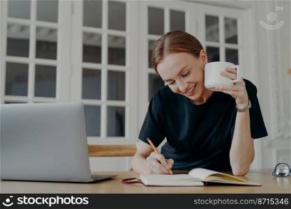 Smiling young woman writing ideas, goals, taking notes, sitting at desk with laptop. Happy freelance female writes in notebook, planning day schedule in the workplace, drinking coffee at home.. Smiling woman writing, taking notes in notebook, planning day schedule, sitting at desk with laptop