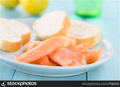 Smoked salmon slices with baguette on blue plate, lemon and glass in the back, photographed on blue wood (Selective Focus, Focus one third into the image) . Smoked Salmon Slices
