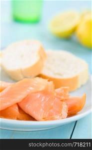 Smoked salmon slices with baguette on blue plate, lemon and glass in the back, photographed on blue wood (Selective Focus, Focus one third into the image) . Smoked Salmon Slices