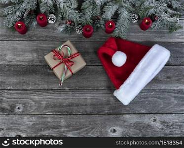 Snow covered evergreen branches with bright red burning candles, giftbox and a Santa cap, on faded wooden planks for a merry Christmas or happy New Year holiday celebration concept