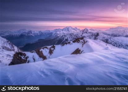 Snow covered mountains and colorful purple sky with clouds at sunset in winter. Beautiful wintry landscape with snowy rocks and hills at dusk. Scenery with alps at frosty evening. Alpine mountains
