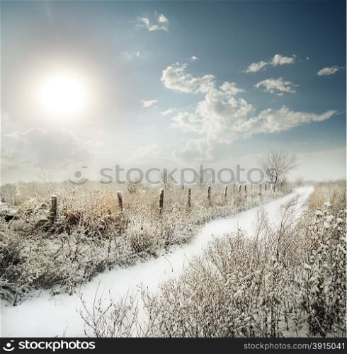 Snow-covered road past the fence in a field under a cloudy sky