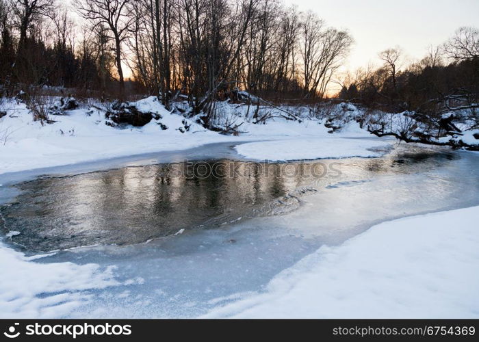 snow riverbank of forest stream at winter sunset