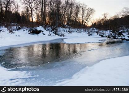 snow riverbank of forest stream at winter sunset