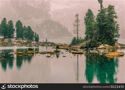 Snowfall on the mountain lake. Reflection of rocks and trees in the water surface. First snow in the mountains.
