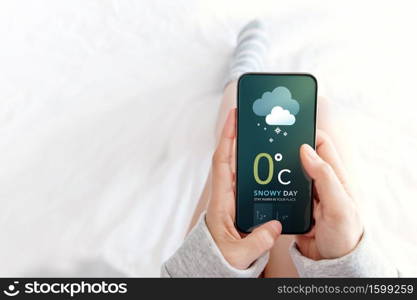 Snowy Day Concept. Cold Weather in Winter Season. Female Lying in Bed while Using Mobile Phone to see Weather Forecast on Screen. Top View