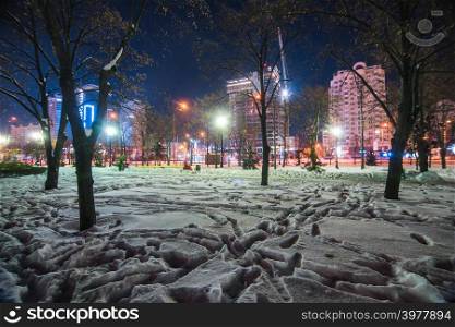 Snowy footprints in night illuminated park with default architecture on background