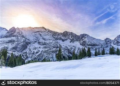 Snowy mountain peaks - Lovely winter scenery with the rocky peaks of the Austrian Alps mountains, their evergreen fir forests and the pastures covered in snow, everything warmed up by a colorful sun.