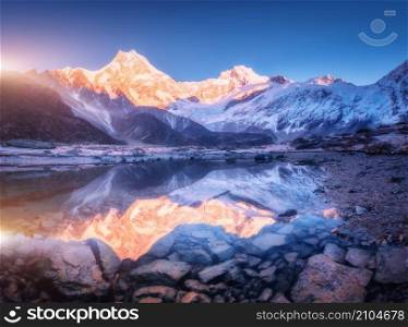 Snowy mountain with illuminated peaks is reflected in beautiful lake at sunrise. Landscape with lighted rocks, violet sky, pond, stones in water at dawn. Winter Himalayan mountains in Nepal. Nature