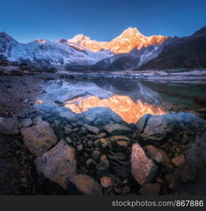 Snowy mountain with illuminated peaks is reflected in beautiful lake at sunrise. Panoramic landscape with lighted rocks, blue sky, pond, stones in water at dawn. Himalayan mountains in Nepal. Nature