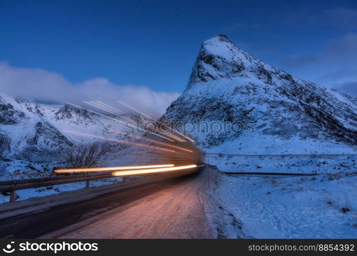 Snowy mountains and blurred car headlights on the road in winter at night. Lofoten Islands, Norway. Beautiful landscape with rock in snow, clouds, blue sky and road at twilight. Light trails. Nature