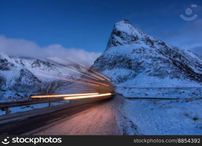 Snowy mountains and blurred car headlights on the road in winter at night. Lofoten Islands, Norway. Beautiful landscape with rock in snow, clouds, blue sky and road at twilight. Light trails. Nature