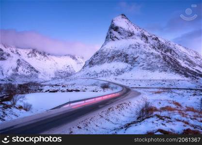 Snowy mountains and red blurred car headlights on the road in winter at dusk. Lofoten Islands, Norway. Landscape with rock in snow, low violet clouds, blue sky and rural road at twilight. Nature